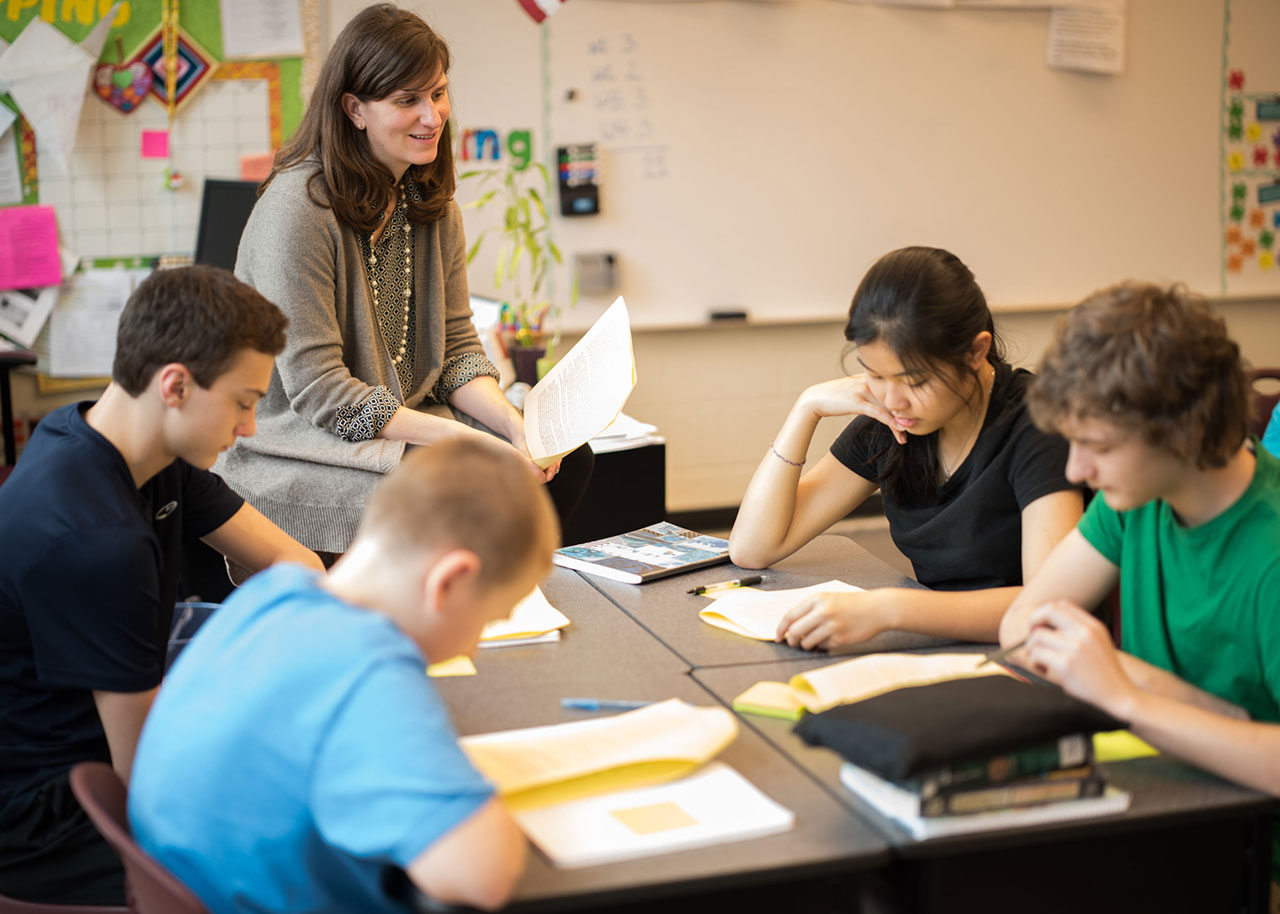 Teacher sits on desk at front of classroom as middle school students read at a table.
