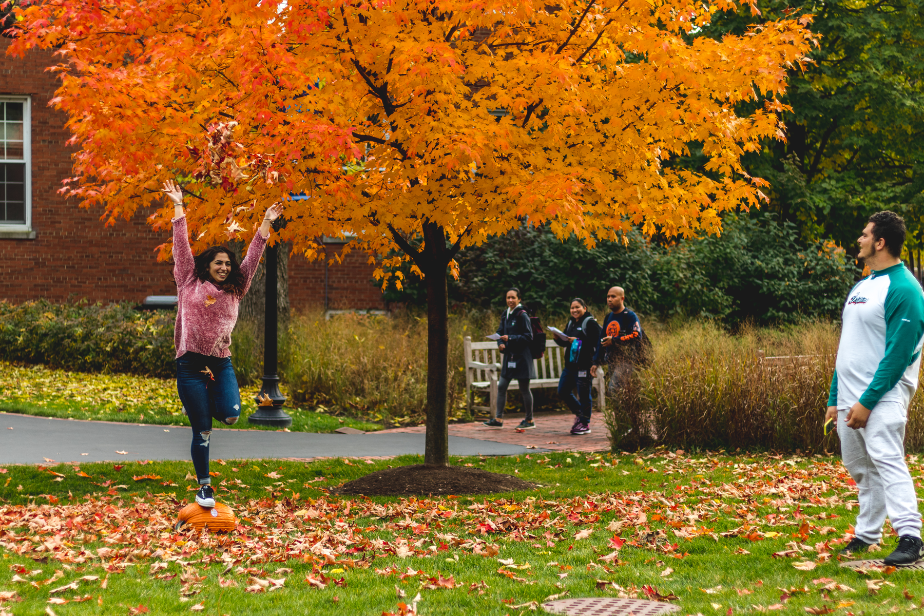 Female student balances on a pumpkin under tree with bright orange leaves. Other students stroll across the greenspace. Campus reopens for the fall 2020 semester, following closure due to COVID-19 concerns.