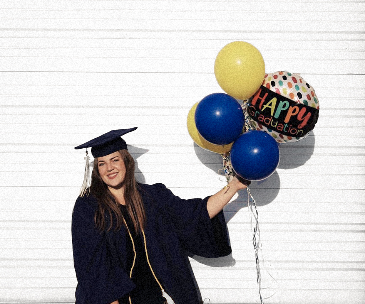 Female student in graduation robe holds blue and yellow balloons.