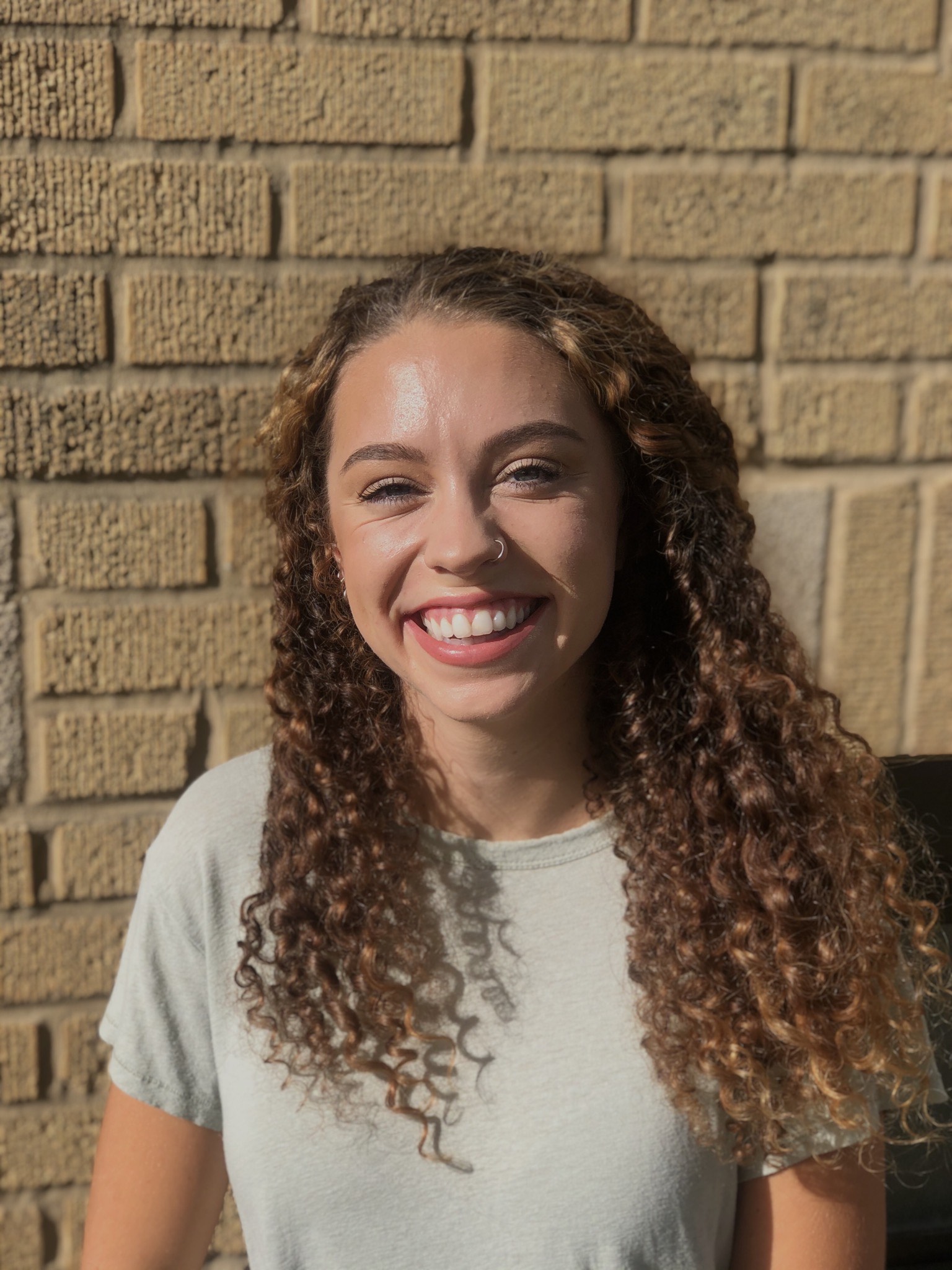 Student with curly brown hair and gray t-shirt stands in front of brick wall.