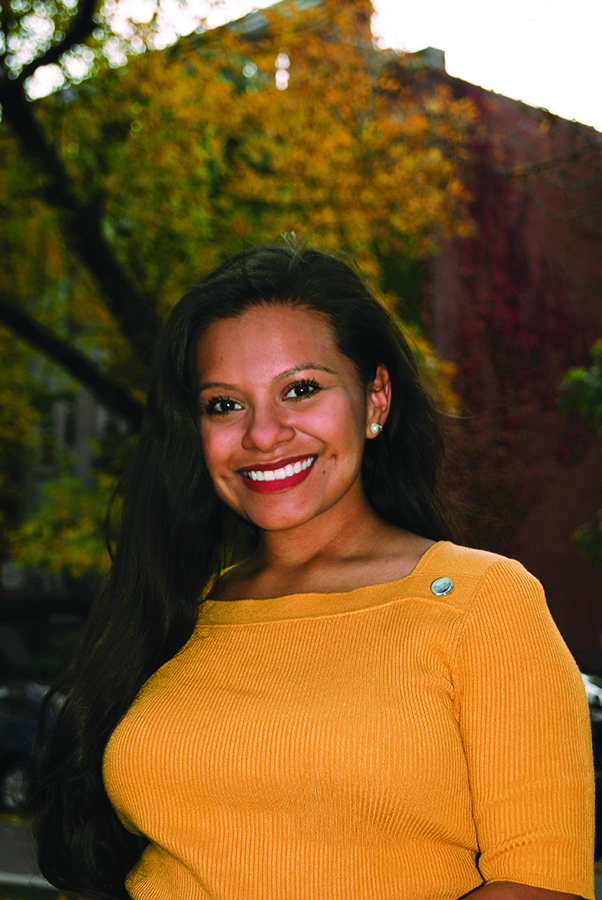 Woman in yellow shirt smiles in front of tree and brick building