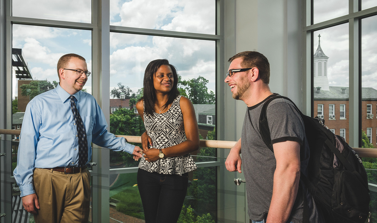 Professor and two students stand chatting in front of full length windows overlooking campus.