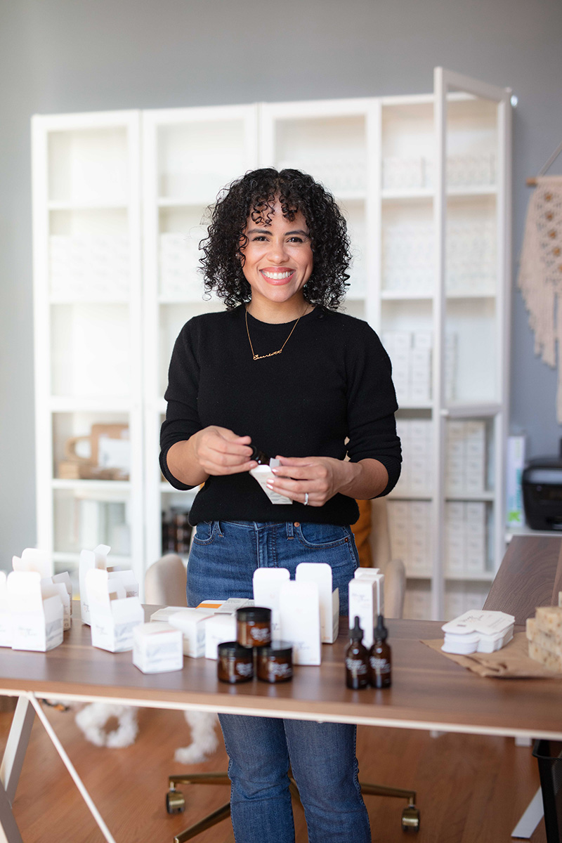 Woman stands in sunlit workspace behind a table spread with personal care products.